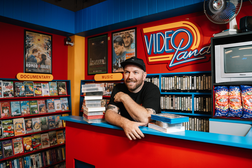 Callum Preston behind the counter of his replica 1990s video store, a place of significance during his carefree and joyous teenage years. 