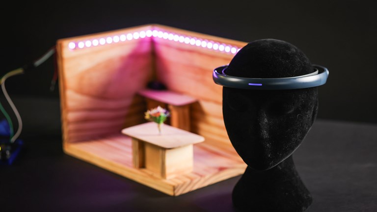Black mannequin head wearing a plastic headband with a purple light. In the background is a scale model room with LED lighting around the walls.