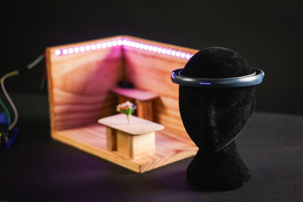 Black mannequin head wearing a plastic headband with a purple light. In the background is a scale model room with LED lighting around the walls.