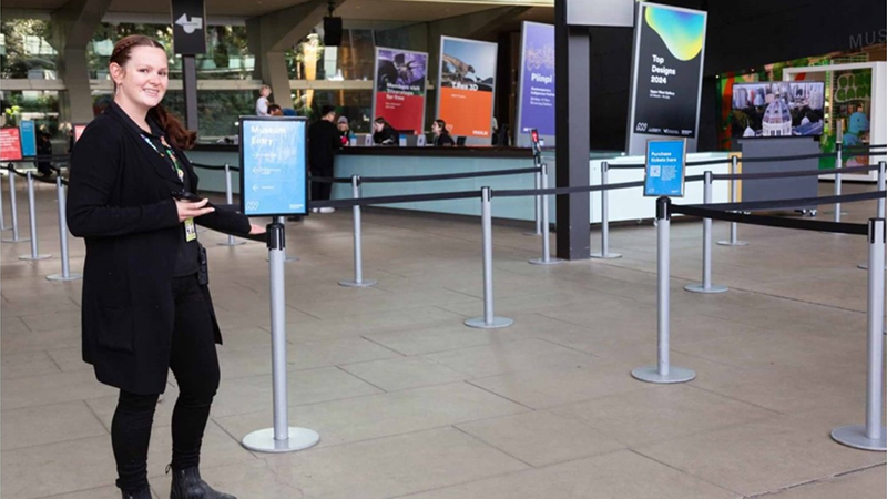A museum staff member, wearing all black clothes and a coloured lanyard standing in the museum foyer.