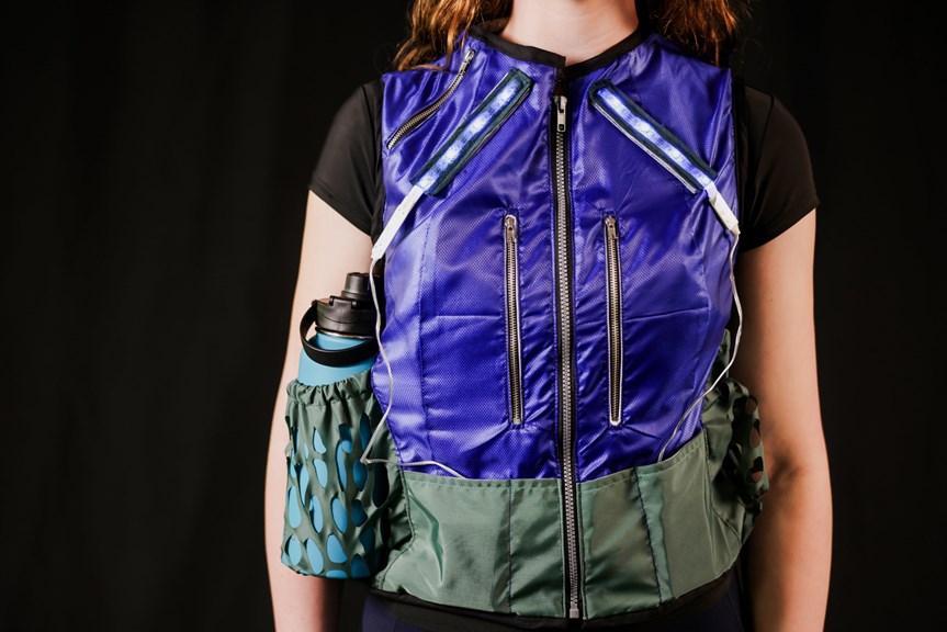 Person wearing a bright blue and green vest with silver zips, LED lights, and a water bottle in the right side pocket.