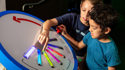 Two children explore an interactive colour exhibit, using a circular panel with glowing coloured tubes arranged like a spectrum. 