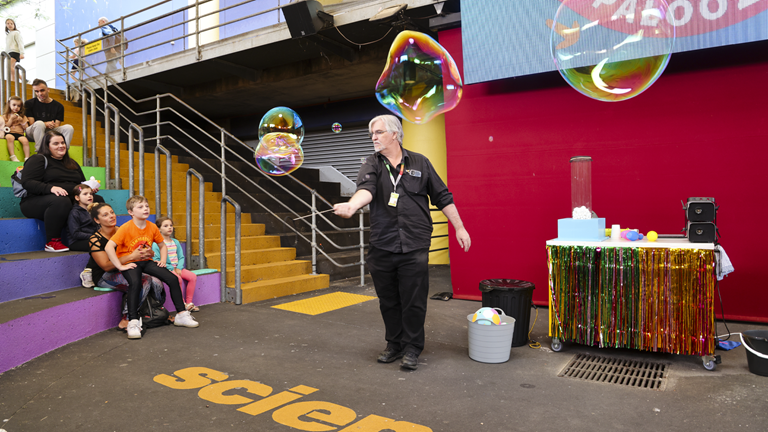 A presenter stands on a stage making large bubbles. They are dressed in all black. There are families looking on from the audience. A screen behind the stage says ‘Air-palooza’ and written on the stage is the beginning of the word ‘Scienceworks’. A variety of other science props are also on stage.