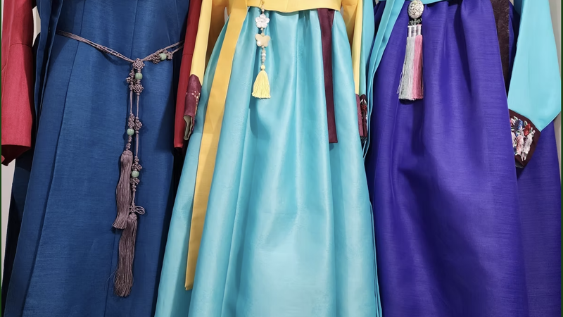 Three traditional Korean garments, one a vest-like robe, and two with a cropped long-sleeve top, empire waist, and balloon skirt, are displayed in a row. Each is a mix of royal gemstone colours like purple, green, peach and aqua.