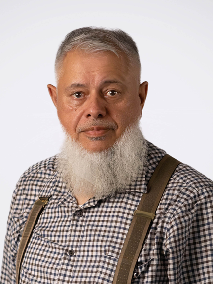 A bearded Indigenous Elder, wearing a checkered shirt and brown suspenders, looks directly into the camera.