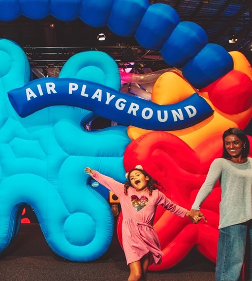 A girl and her carer in front of the Air  Playground exhibition. The girl has her hands are held above her head with excitement.
