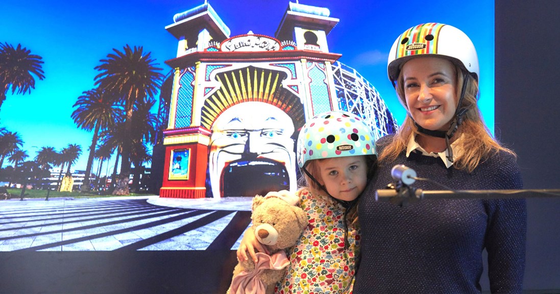 A woman and young girl wear bike helmets and smile for a photo. Behind them is a digital background of Luna Park. 