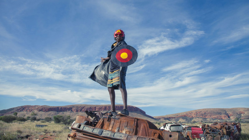 A man wearing a captain America costume standing on a rusted out car in the Australian outback.