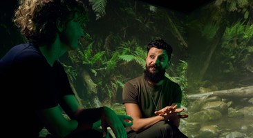 Two people sitting on stools having a conversation. A large image of a forest covers the wall in the background.