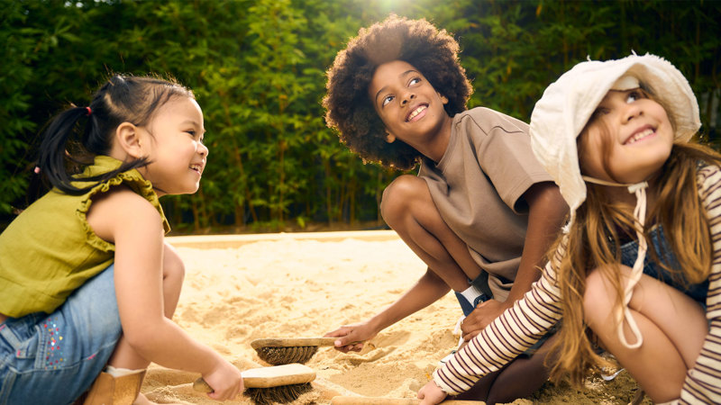 Three small children playing in a sandpit using dustpan brushes to sweep the sand.