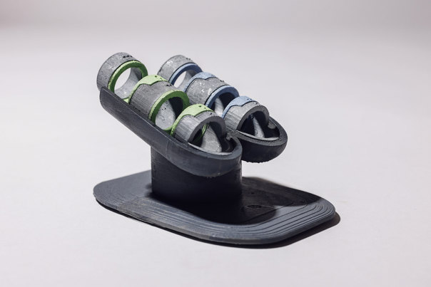3D-printed finger exercise or therapy device with grey cylindrical components featuring green and blue accent rings, displayed on a black base.