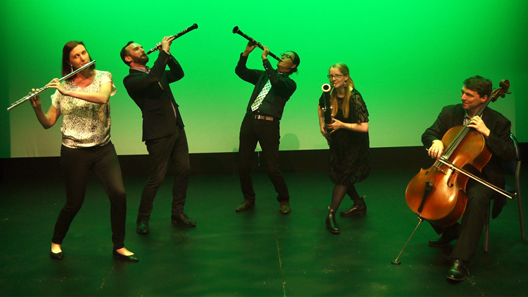 Five Musicians standing in front of a green wall playing the flute, clarinet, oboe basoon and cello