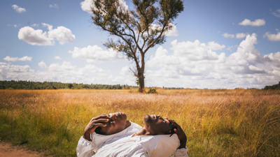 Two men in white hazmat suits, faces exposed, lean back on each other with closed eyes pointing at the sky.