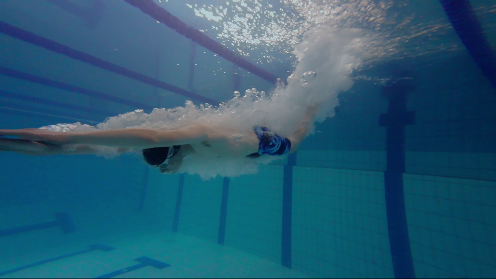 Underwater view of a swimmer wearing goggles and board shorts, diving into a tiled pool.