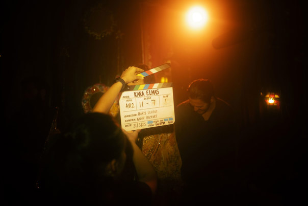 A spotlight shines on a woman opening a clapperboard with the film’s title KARA ELMAS displayed in bold black lettering. In the background you can vaguely see an actress in traditional festive clothing, a man and a blurry yellow backlight. 