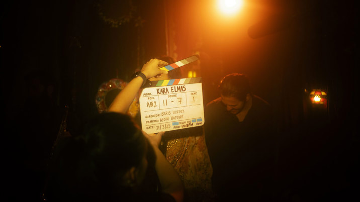 A spotlight shines on a woman opening a clapperboard with the film’s title KARA ELMAS displayed in bold black lettering. In the background you can vaguely see an actress in traditional festive clothing, a man and a blurry yellow backlight. 