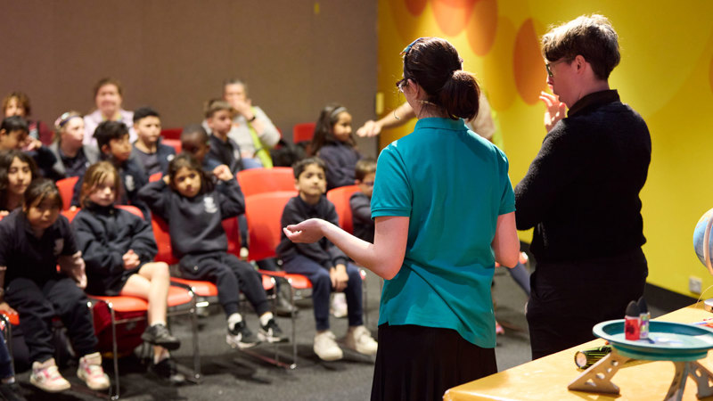 Students sitting in an audience watching a museum staff present a show about rockets at Scienceworks with a demonstration table behind them.  