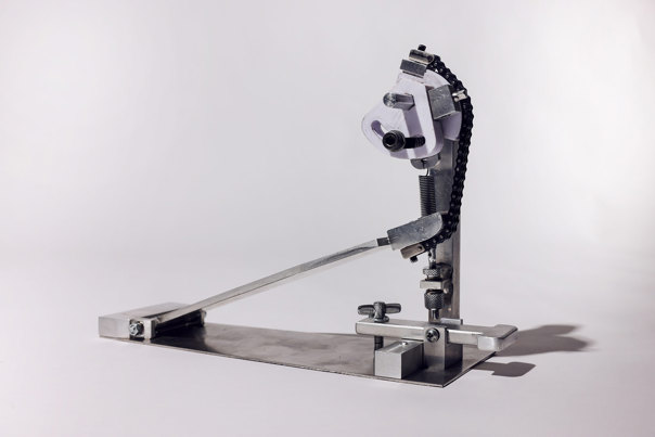 Metal bass-drum pedal mechanism with articulated arm and chain drive system, mounted on rectangular base plate.