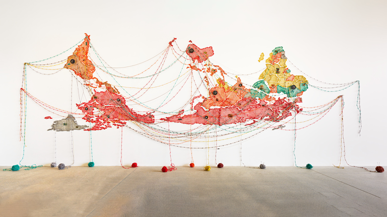 Variously coloured balls of wire stretch between many pins on the wall to image an upside down map of the world before falling to the ground in a ball