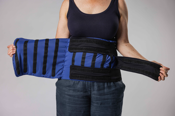 Person in a dark top and trousers holds open a blue and black abdominal support belt with multiple fastenings.