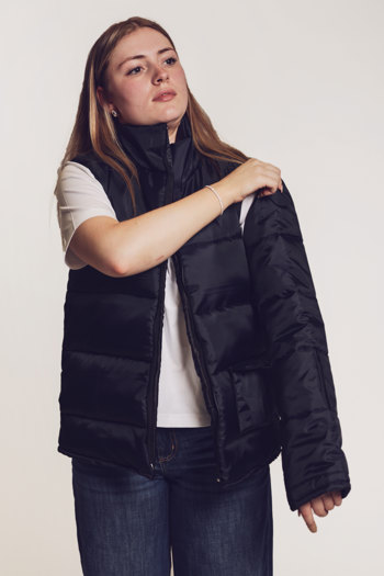 Model standing against a neutral backdrop, wearing jeans, a white T-shirt and a dark-coloured puffer jacket with detachable sleeves. One sleeve has been removed and she is unzipping the other sleeve.