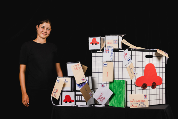 Woman in a black shirt standing next to a display of cube-shaped packages decorated with grid patterns, red car cut-outs, and handwritten tags.