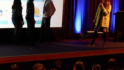 Three students on stage in the Museum Theatre silhouetted in front of a powerpoint about clean energy use, pointing to different years  of technology adoption. To their right, a Museum expert gives a presentation. 