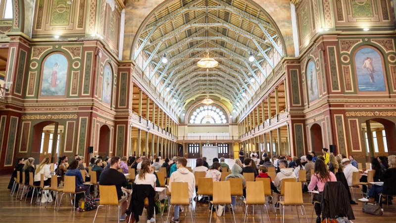 A group of budding artists sis on chairs under the dome of the Royal Exhibition Building for a drawing class.