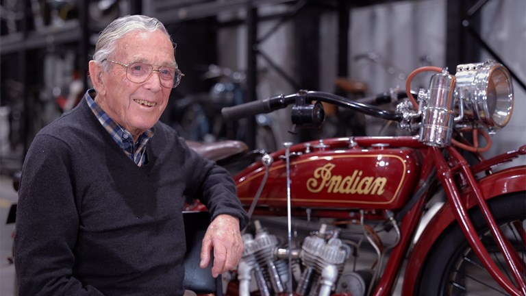 An elderly man smiling while sitting next to a motorcycle