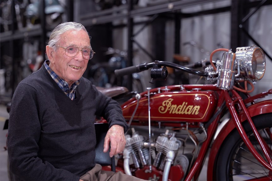 An elderly man smiling while sitting next to a motorcycle