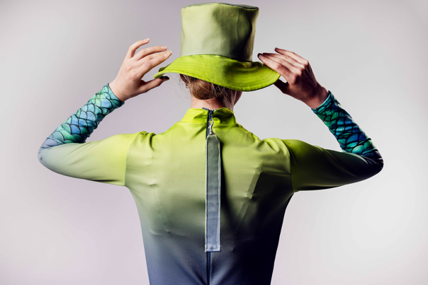 Person seen from behind wears a green long-sleeved rash top with fish-scale patterned sleeves and a matching bucket hat.