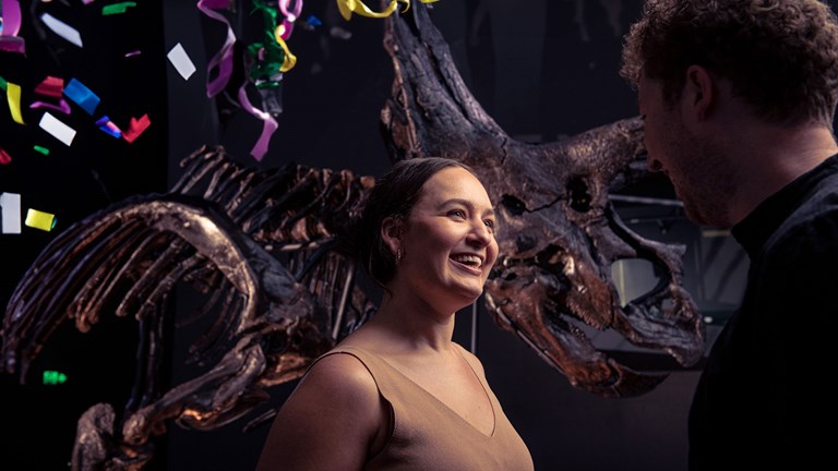 Two people stand talking and laughing in front of Horridus, the Triceratops amidst party streamers
