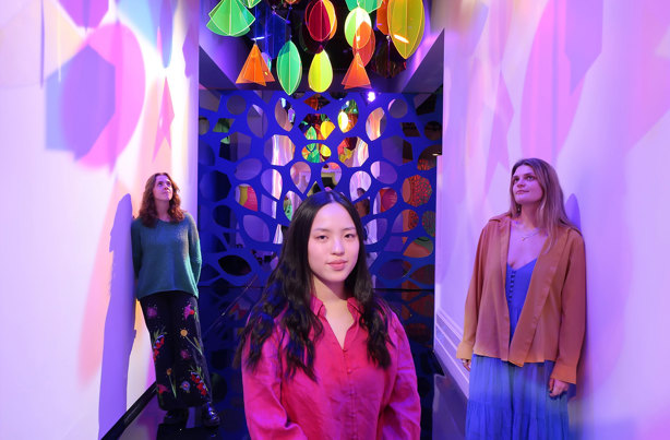Looking down a hallway with a mosaic of mirrors at the end, two women lean on opposite white walls looking upwards and a third woman stands in the middle looking directly at the camera. Hanging from the ceiling are rainbow-coloured, translucent geometric shapes and spotlights, casting colourful shadows on the walls.
