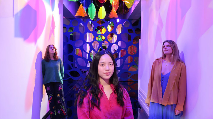 Looking down a hallway with a mosaic of mirrors at the end, two women lean on opposite white walls looking upwards and a third woman stands in the middle looking directly at the camera. Hanging from the ceiling are rainbow-coloured, translucent geometric shapes and spotlights, casting colourful shadows on the walls.