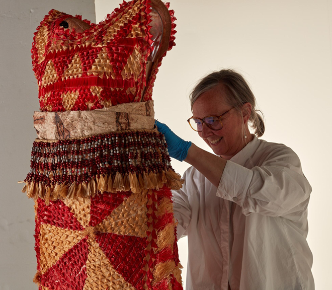 A blonde woman in a white shirt, and wearing glasses, pins a beaded red and yellow dress to a mannequin.