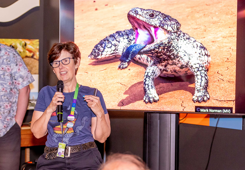 A woman in front of a screen displaying a blue-tongued lizard gives a talk.
