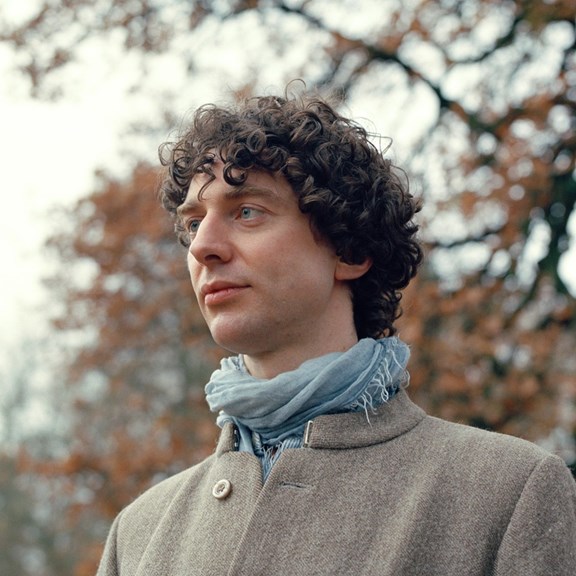 A head shot of a man with short curly hair. He is outdoors wearing a blue scarf and fawn coat. 