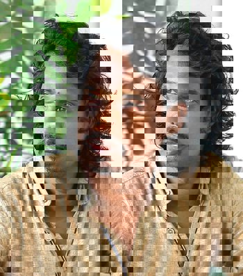 An Indian man with shoulder length curly hair, a mustache and goatee, and wearing a sand coloured collared shirt, looks to the side. 