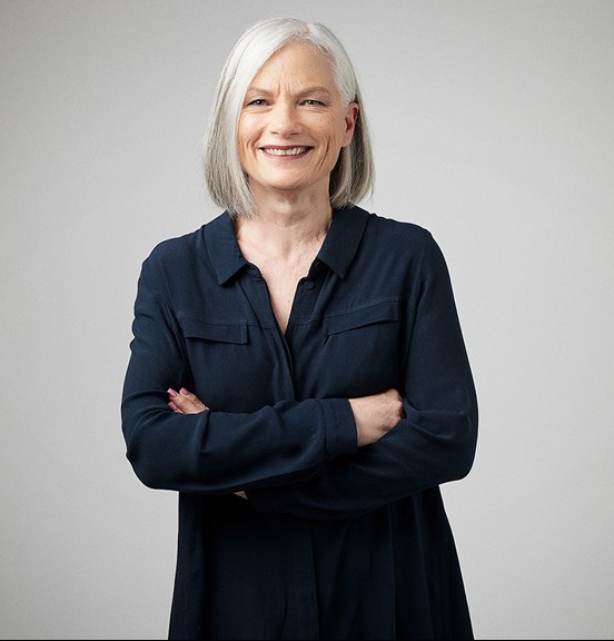 A woman with a grey bob and wearing a black shirt, crosses her arms and smile directly at the camera.