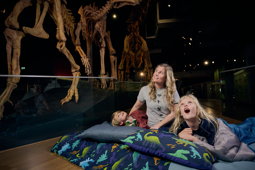 Children and their guardian in sleeping bags beneath giant dinosaur skeletons.