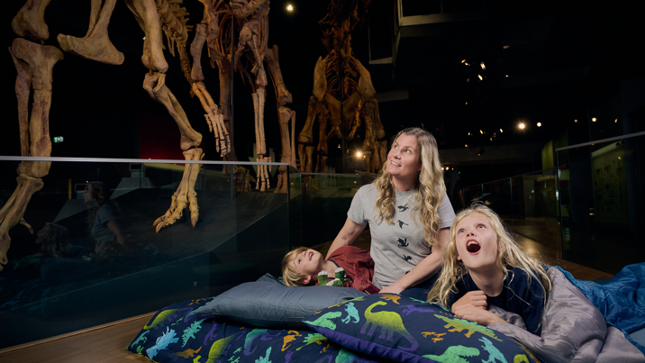 Children and their guardian in sleeping bags beneath giant dinosaur skeletons.