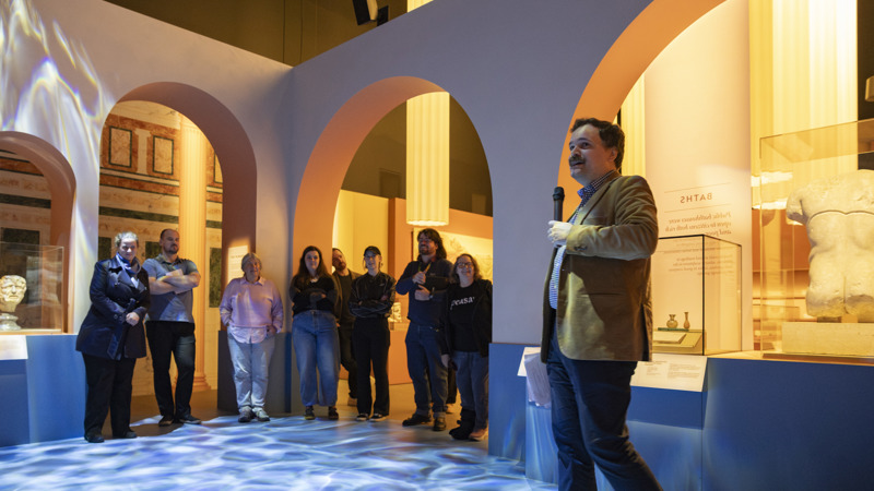 Man in a tweed jacket with a microphone delivering a tour of Rome, an exhibition at Melbourne Museum. Audience listening in the background.