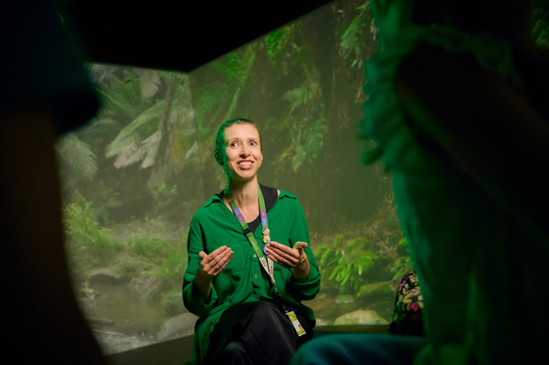 Climate Science Educator Elke Barczak sitting speaking to students, in front of a projected forest scene background.  