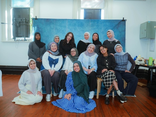 13 members of An.Other Collective smiling at the camera in a room with a blue photo backdrop. 