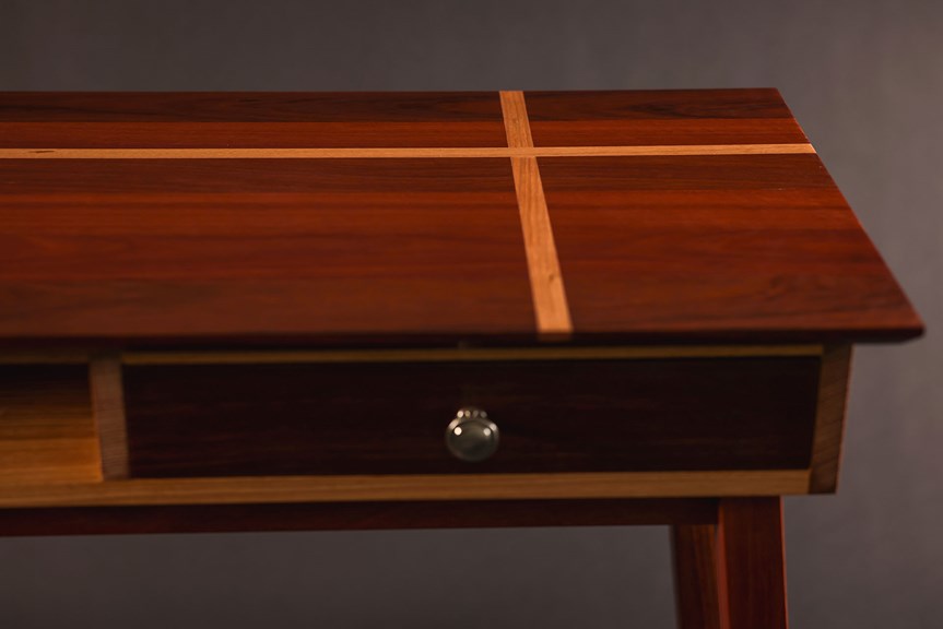 Close-up view of a wooden table that features a cross design on its top and an inset drawer with a silver handle.