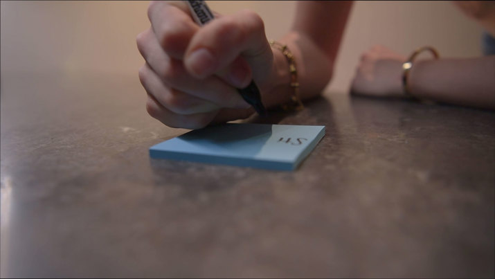 Close-up of a hand writing with a sharpie onto a blue sticky note.