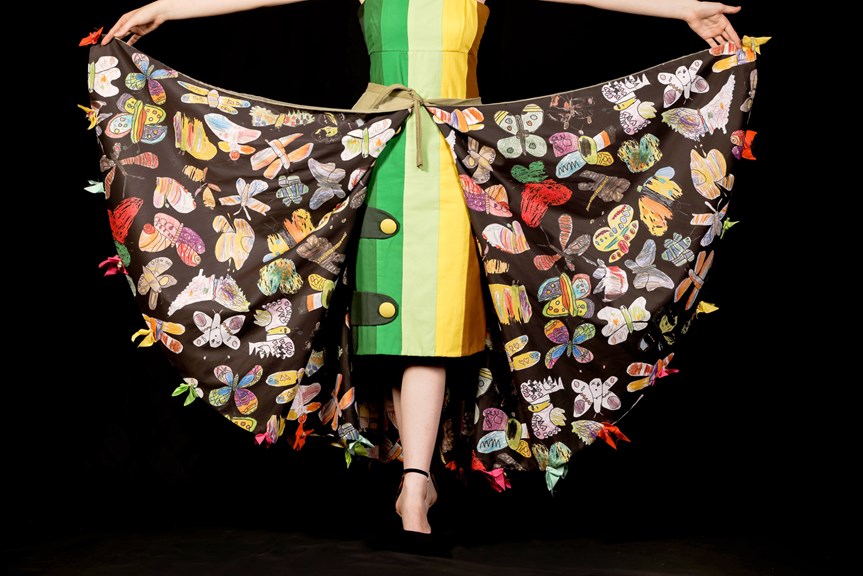 Person wears a cape adorned with butterfly illustrations. A green, yellow and brown shift dress is worn underneath.