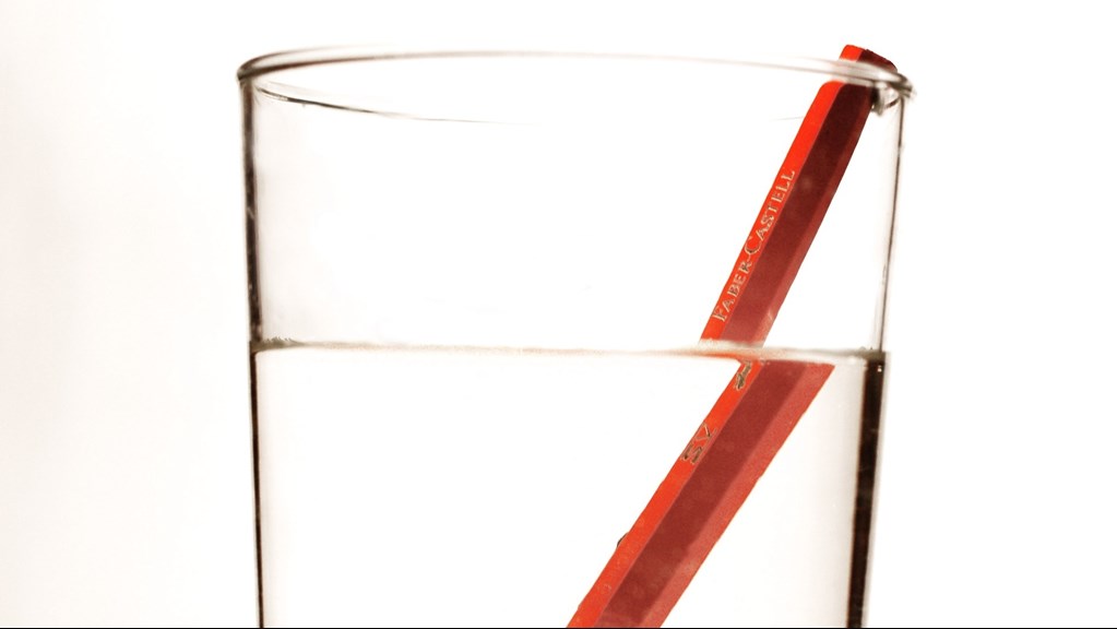 A red pencil has been placed inside a glass of water, as a result of refraction it appears to be horizontally broken.