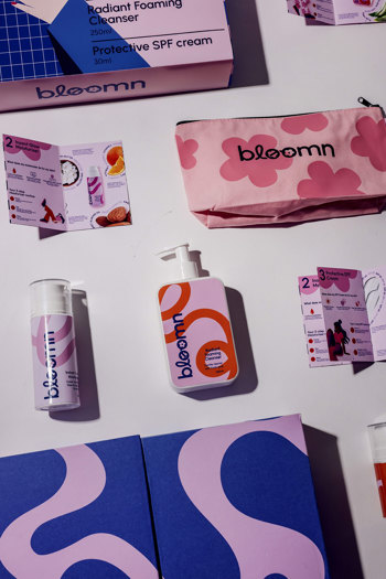 Flat lay of Bloomn skincare product packaging including pump bottles, pink floral cosmetic bag, product information cards, and boxes in pink, blue, lilac and orange colour scheme with bold graphic patterns.