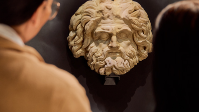 A couple people looking at a large stone statue head of a roman god in ROME: Empire, Power, People exhibition in Melbourne Museum.